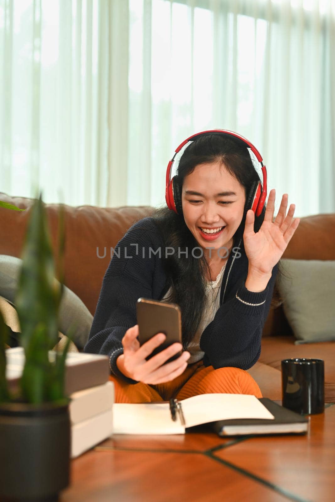 Pretty woman wearing headphone and making video call on her smart phone, relaxing on couch at home by prathanchorruangsak