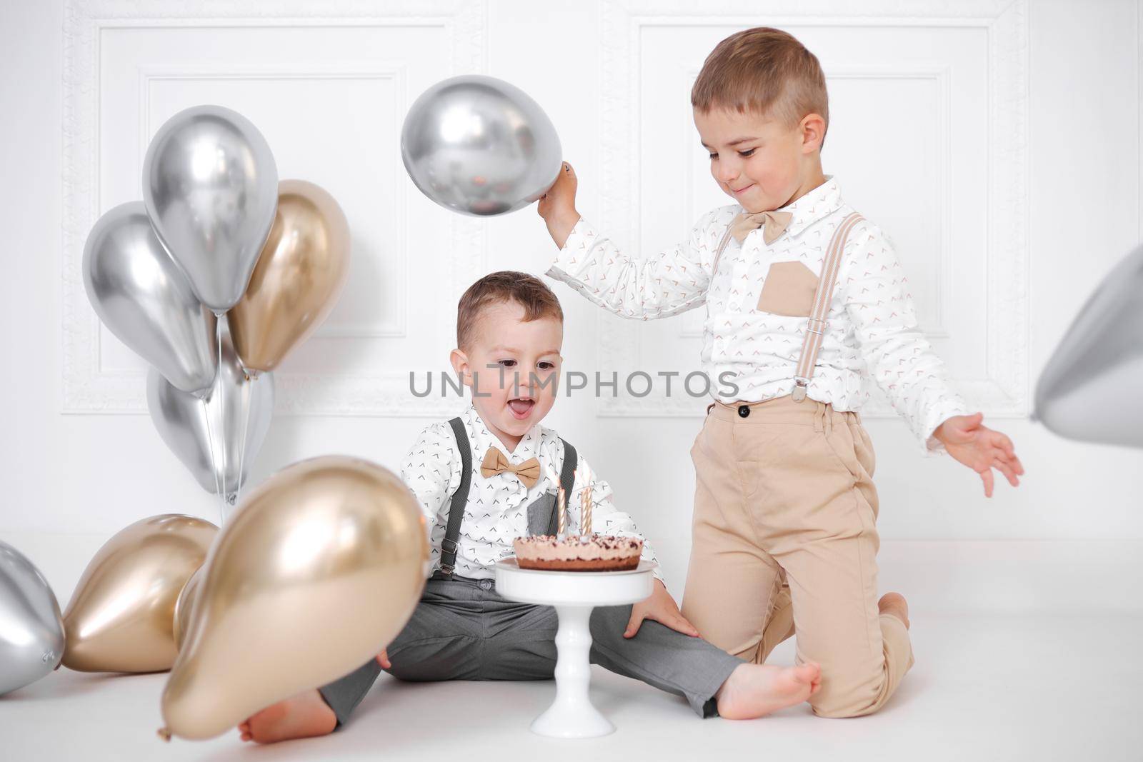 Two boys celebrating birthday, children have a B-day party. Birthday cake with candles and balloons. Happy kids, celebration, white minimalist interior