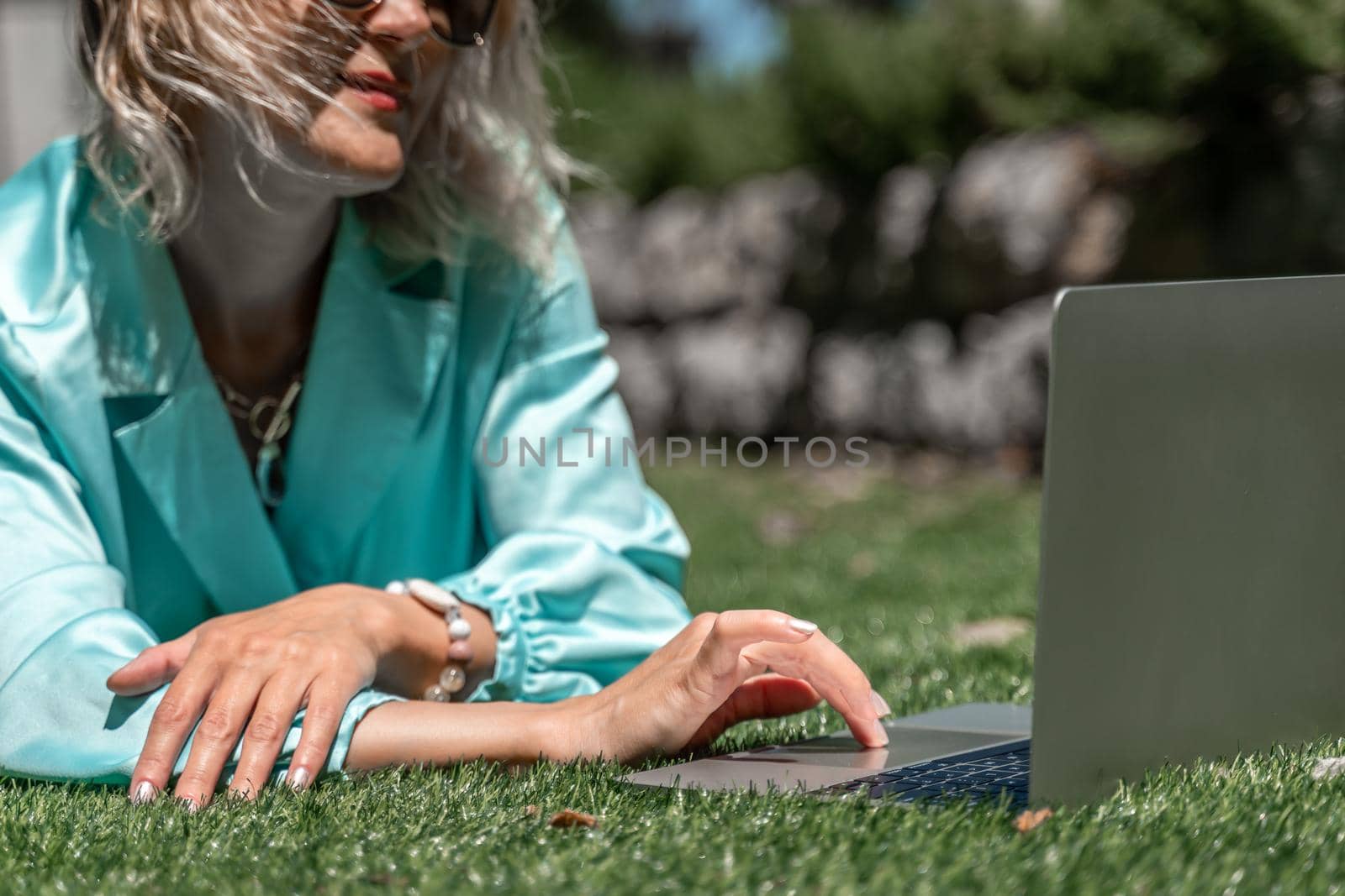 a young beautiful woman with blond curly hair in glasses and a blue dress sits on the grass in nature and uses a laptop, by Matiunina
