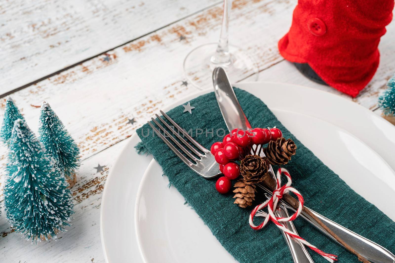 Christmas table setting with white dishware, silverware and red and green decorations on white wooden background. High angle view
