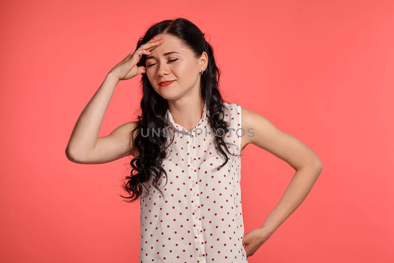 Studio shot of a gorgeous little woman, wearing casual white polka dot blouse. Little brunette female is acting like forget something, posing over a pink background. People and sincere emotions.