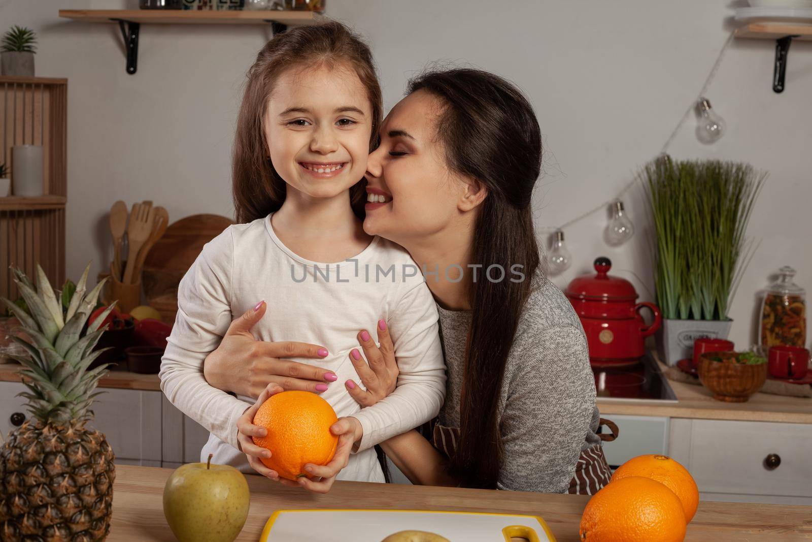 Happy loving family are cooking together. Lovely mommy and her little girl are doing a fruit cutting and hugging at the kitchen, against a white wall with shelves and bulbs on it. Homemade food and little helper.