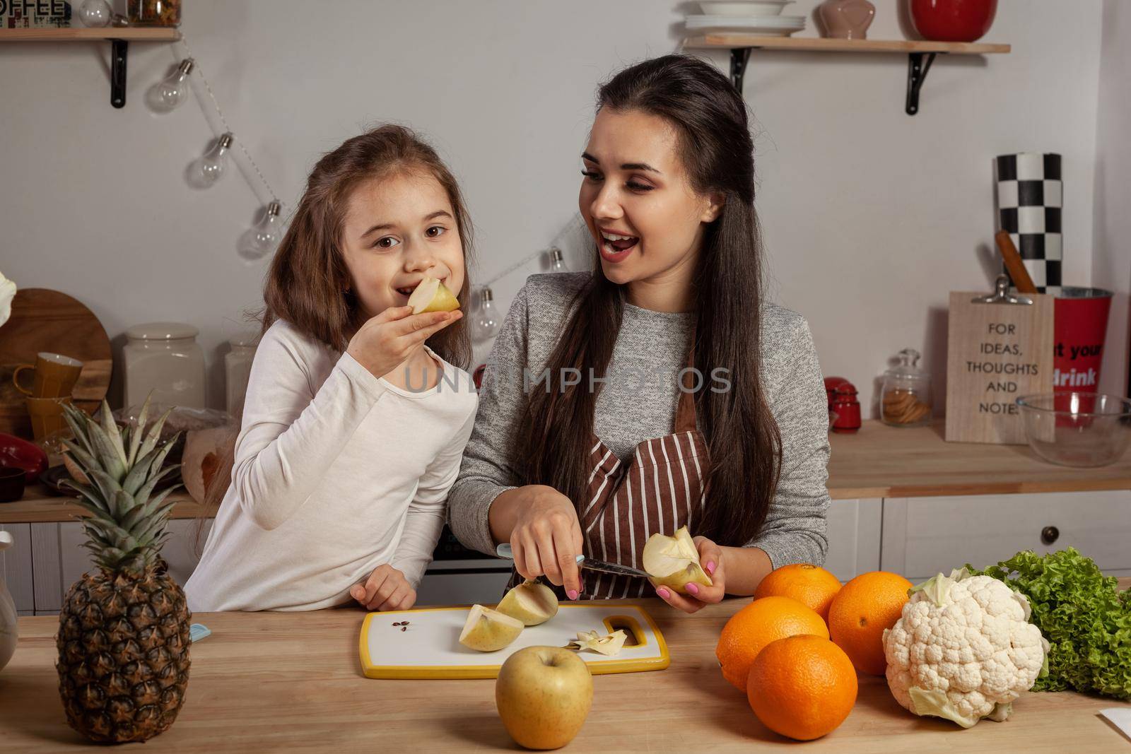Happy loving family are cooking together. Cute mother and her little princess are doing a fruit cutting and tasting an apple at the kitchen, against a white wall with shelves and bulbs on it. Homemade food and little helper.