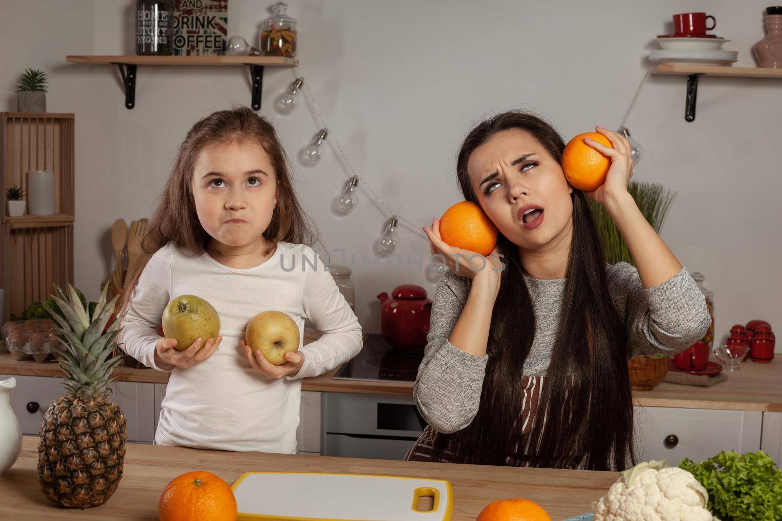 Happy loving family are cooking together. Elegant mother and her daughter are doing a fruit cutting and fooling around at the kitchen, against a white wall with shelves and bulbs on it. Homemade food and little helper.
