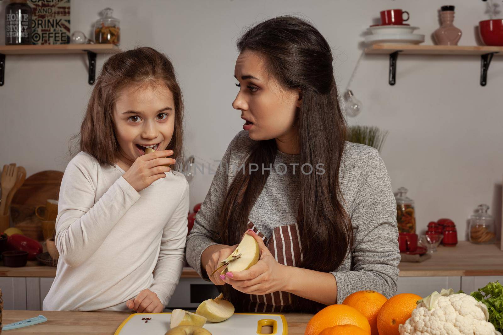 Happy loving family are cooking together. Gorgeous mom and her kid are doing a fruit cutting and tasting an apple at the kitchen, against a white wall with shelves and bulbs on it. Homemade food and little helper.