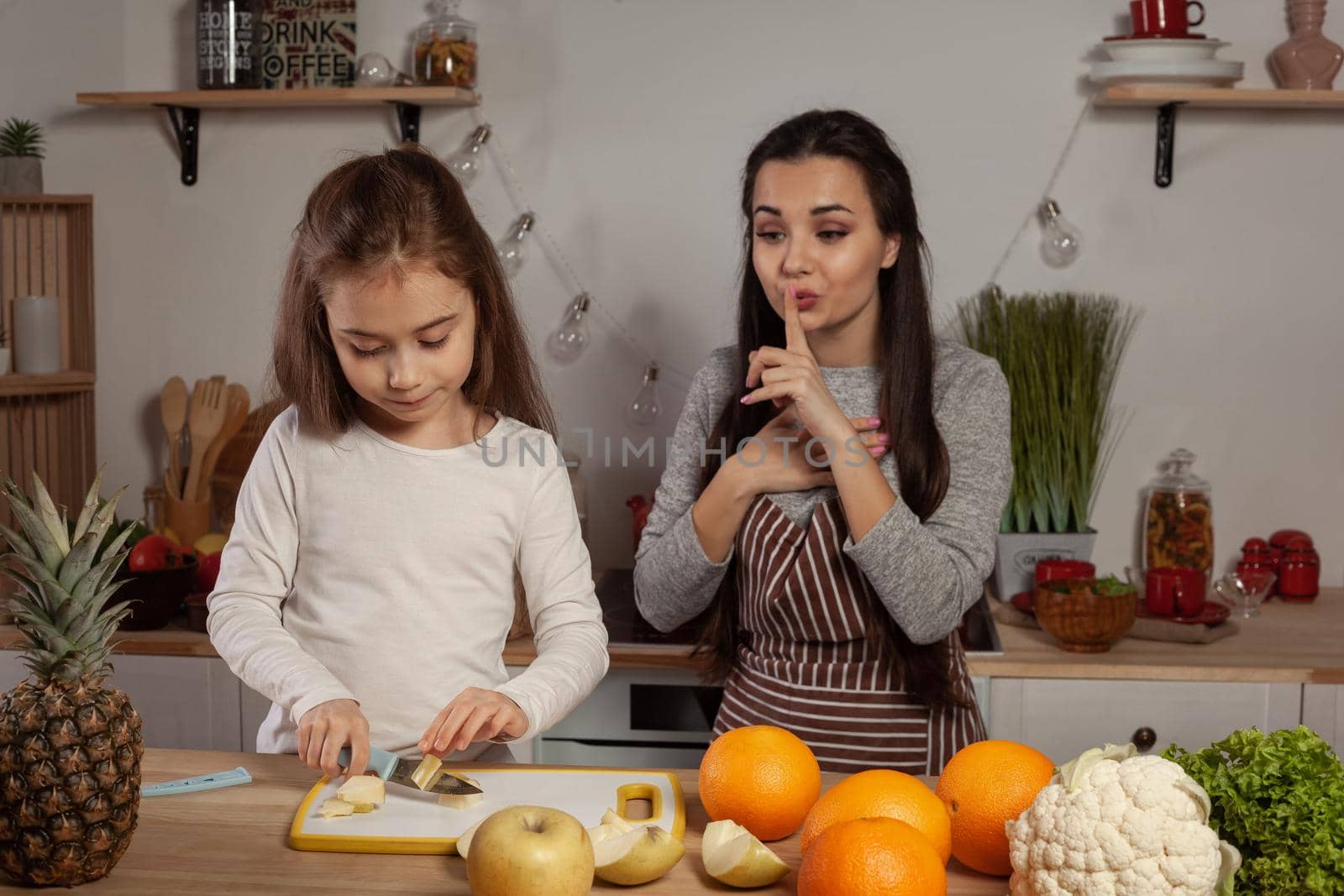 Happy loving family are cooking together. Pretty mum and her child are doing a fruit cutting and mom is admiring about her offspring at the kitchen, against a white wall with shelves and bulbs on it. Homemade food and little helper.