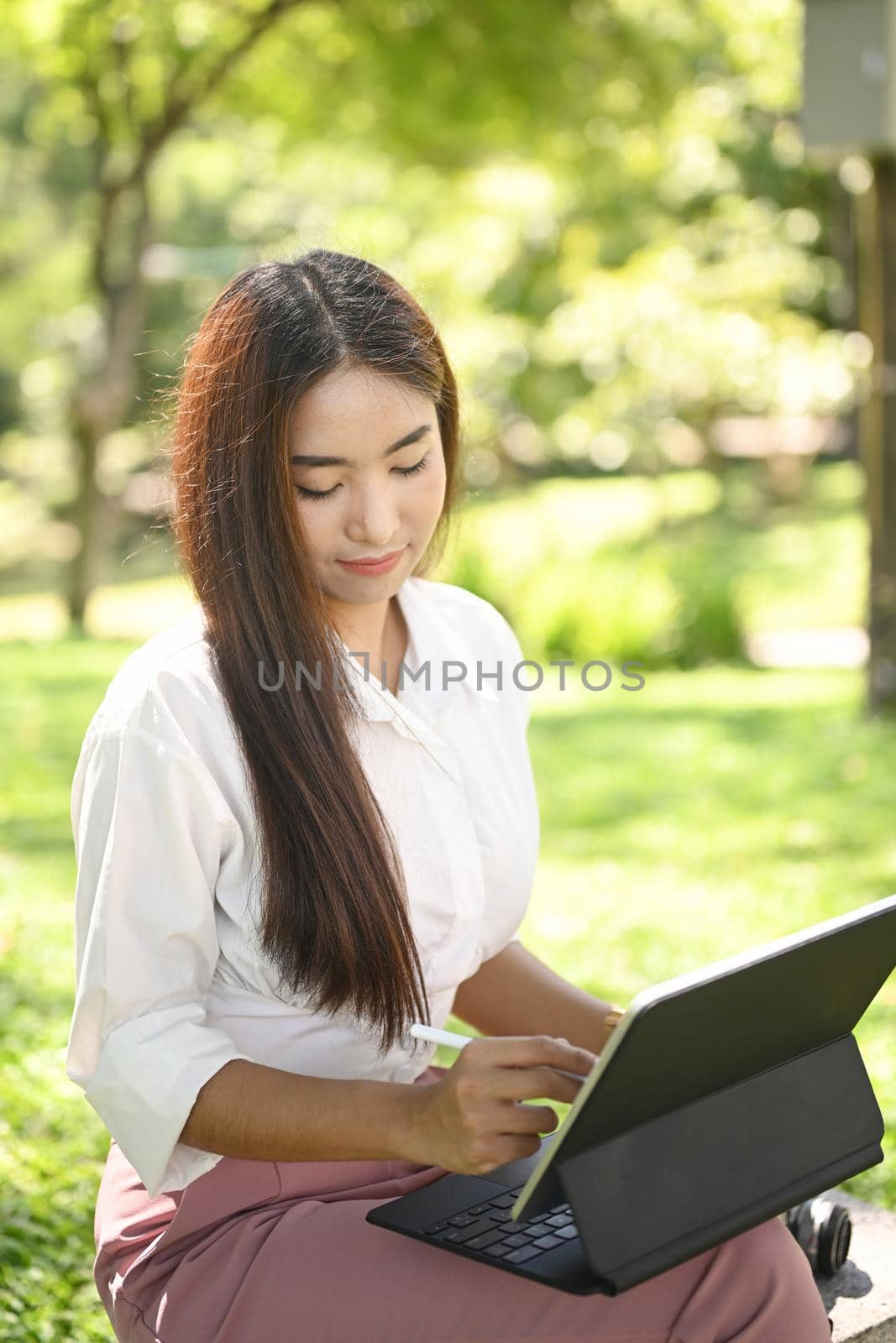 Beautiful Asian woman sitting on bench in the park and working with computer tablet.