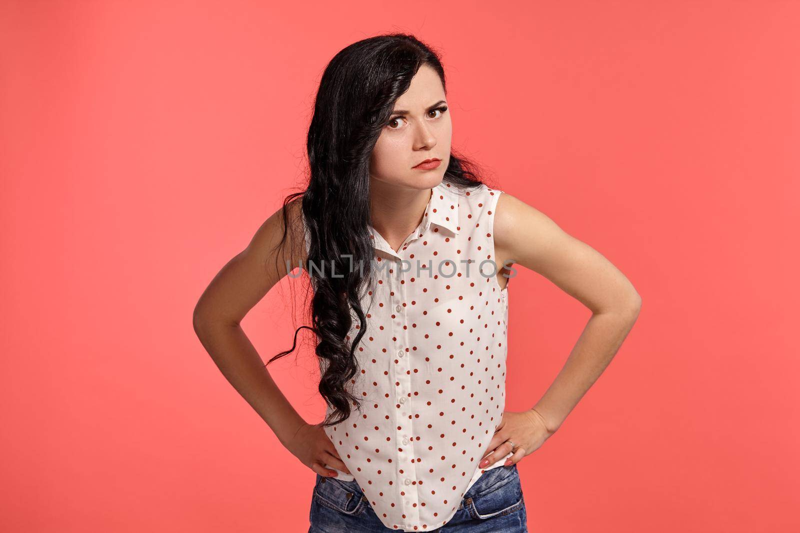 Studio shot of a good-looking little woman, wearing casual white polka dot blouse. Little brunette female is acting like suspect someone, posing over a pink background. People and sincere emotions.