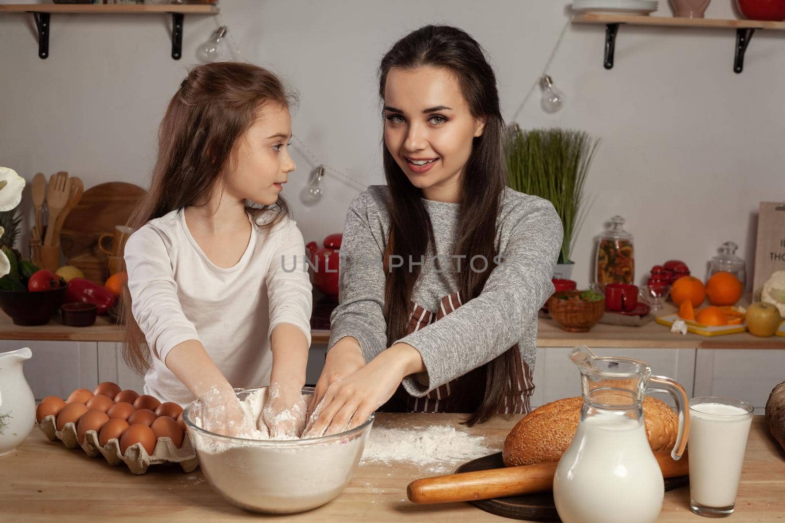 Happy loving family are preparing pastries together. Gorgeous mommy and her little girl are touching a flour and having fun at the kitchen, against a white wall with shelves and bulbs on it. Homemade food and little helper.