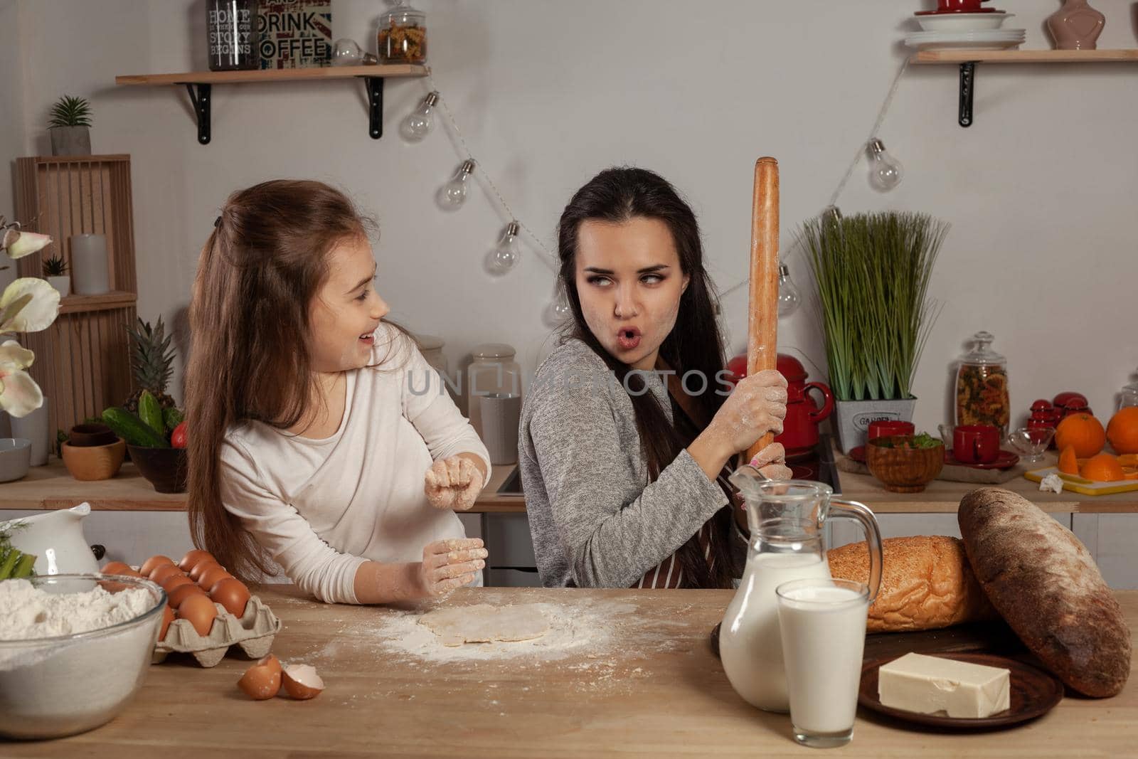 Happy loving family are preparing pastries together. Beautiful mother and her daughter are baking a bread and are fooling around at the kitchen, against a white wall with shelves and bulbs on it. Homemade food and little helper.