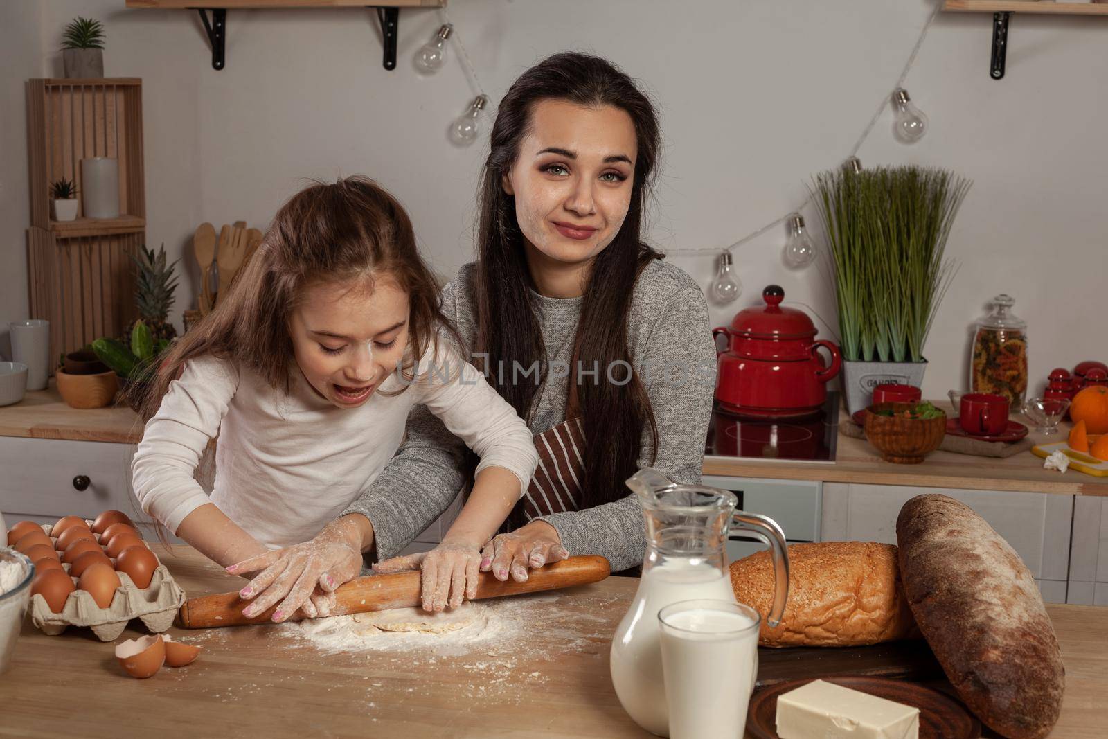 Happy loving family are preparing pastries together. Beautiful mother and her little princess are rolling a dough and having fun at the kitchen, against a white wall with shelves and bulbs on it. Homemade food and little helper.