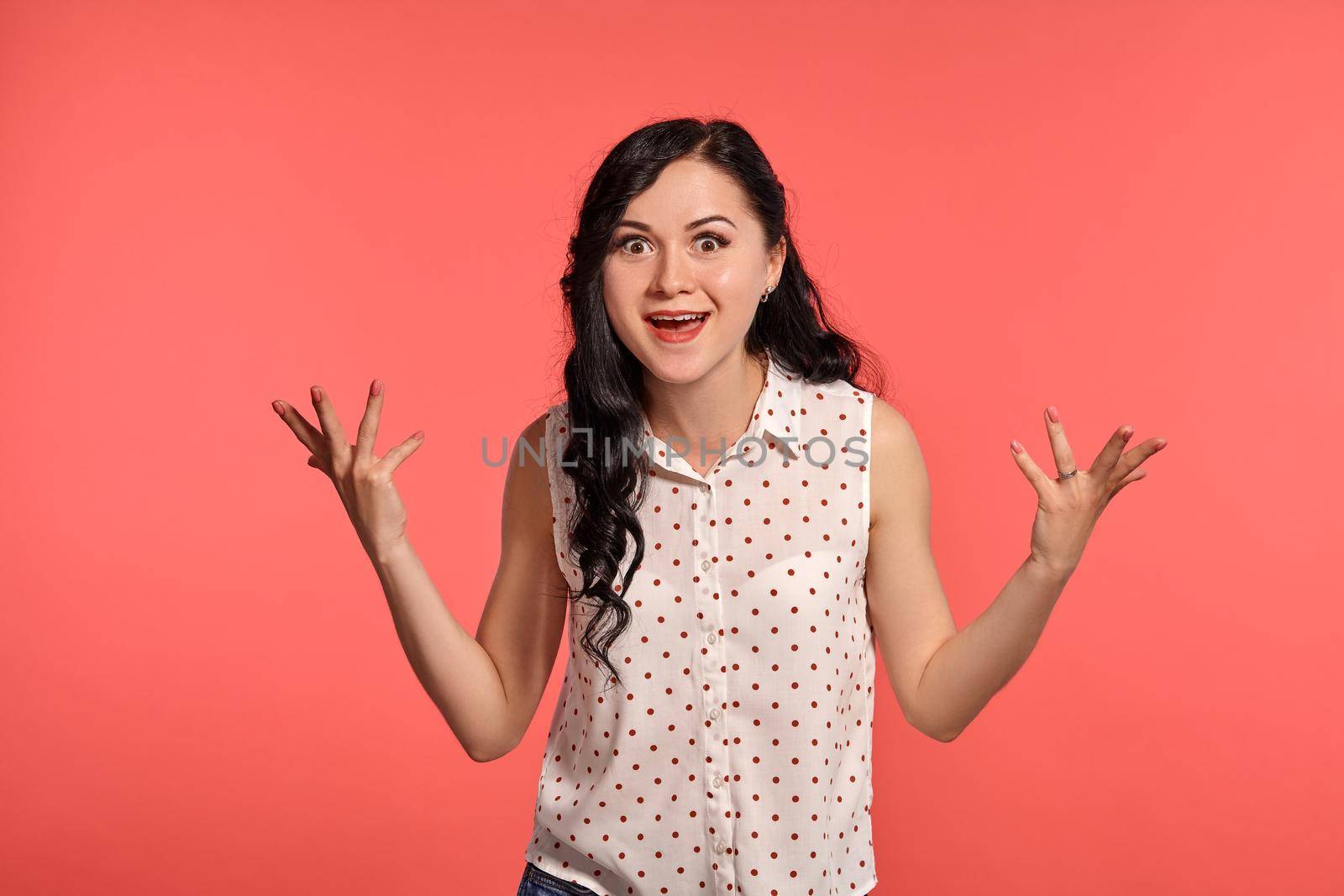 Studio shot of an elegant girl teenager looking surprised, wearing casual white polka dot blouse. Little brunette female feeling happy, posing over a pink background. People and sincere emotions.
