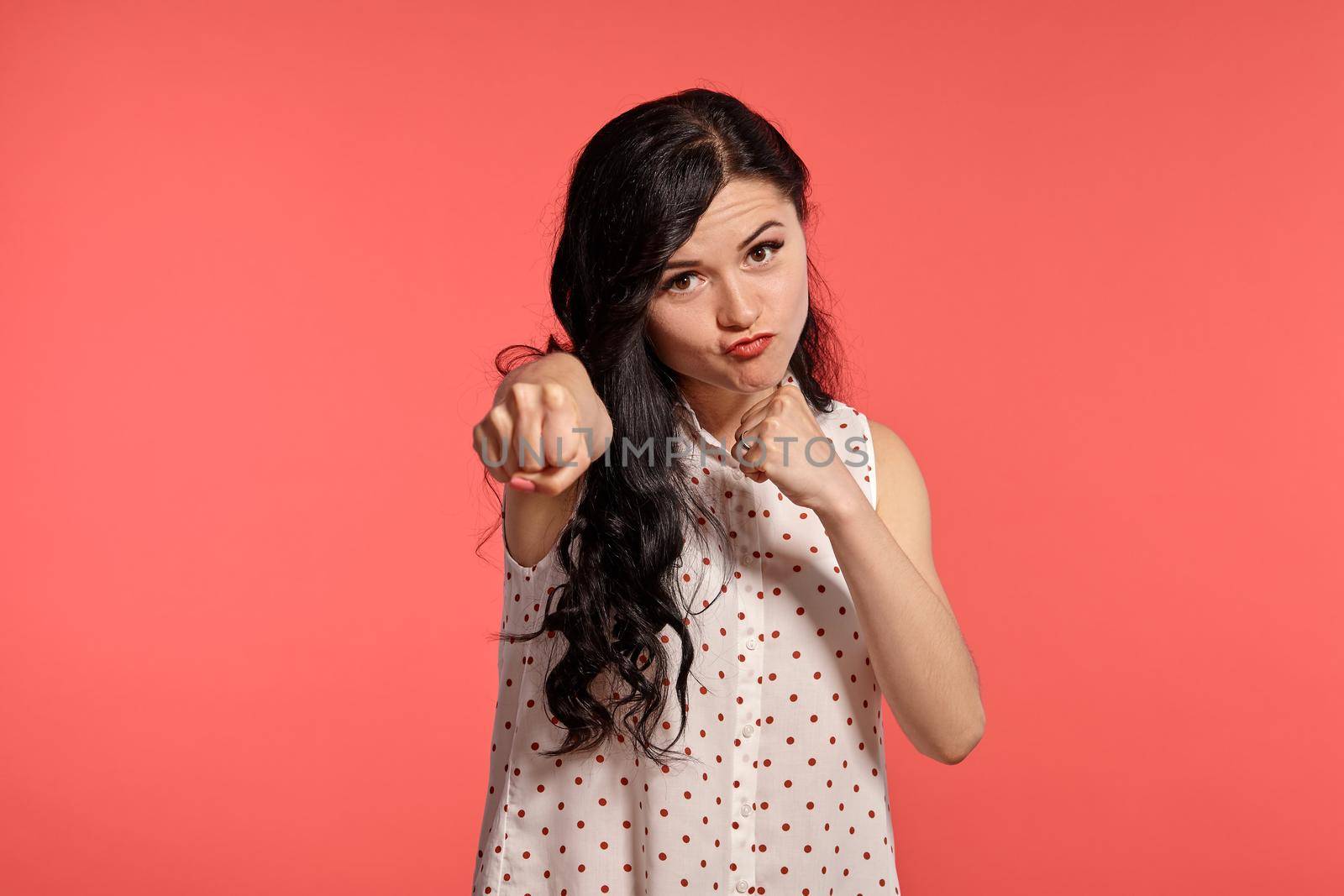 Studio shot of a magnificent teen lady wearing casual white polka dot blouse. Little brunette female is boxing posing over a pink background. People and sincere emotions.