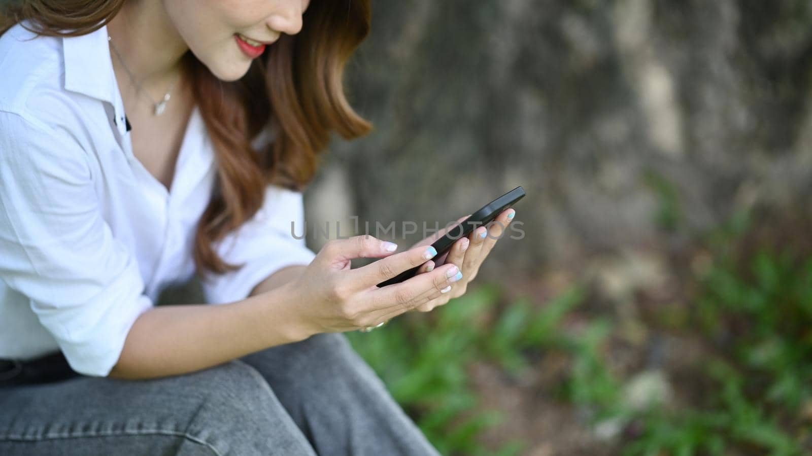 Cropped shot young woman sitting on bench in the nature park and using smart phone. by prathanchorruangsak