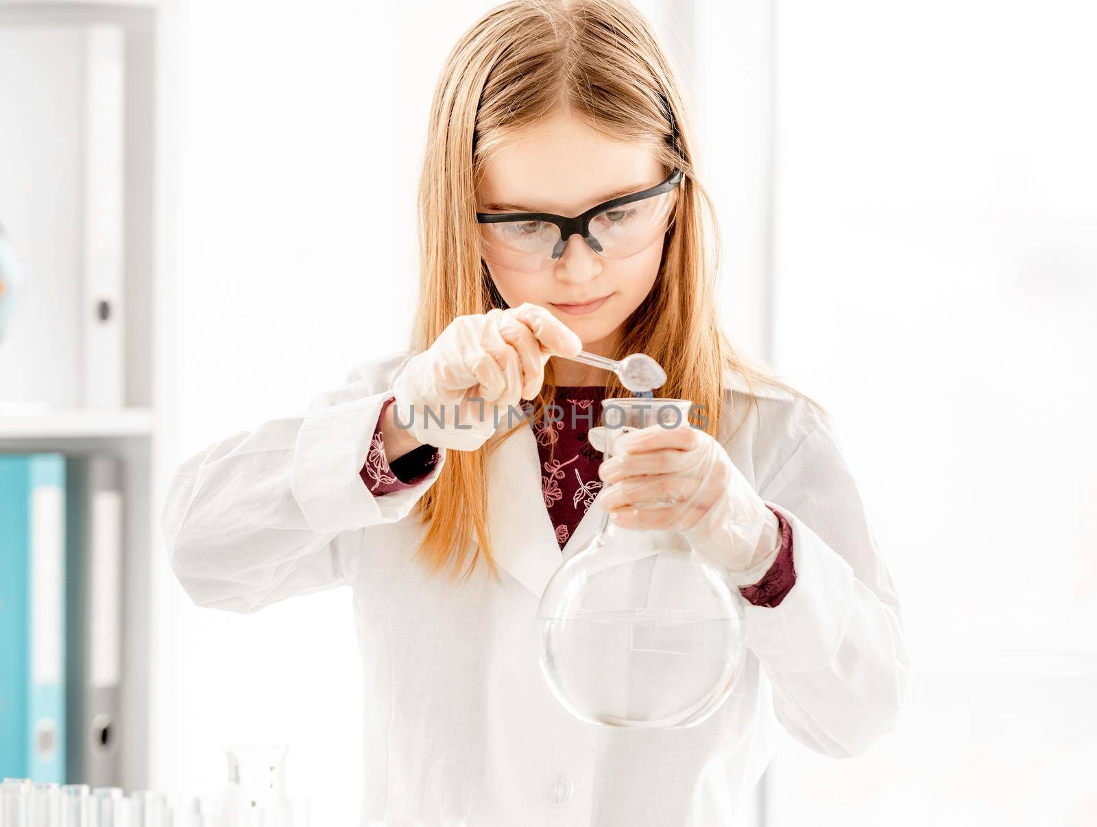 Smart girl doing scientific chemistry experiment wearing protection glasses, holding bottle and measuring ingridients. Schoolgirl with chemical equipment on school lesson