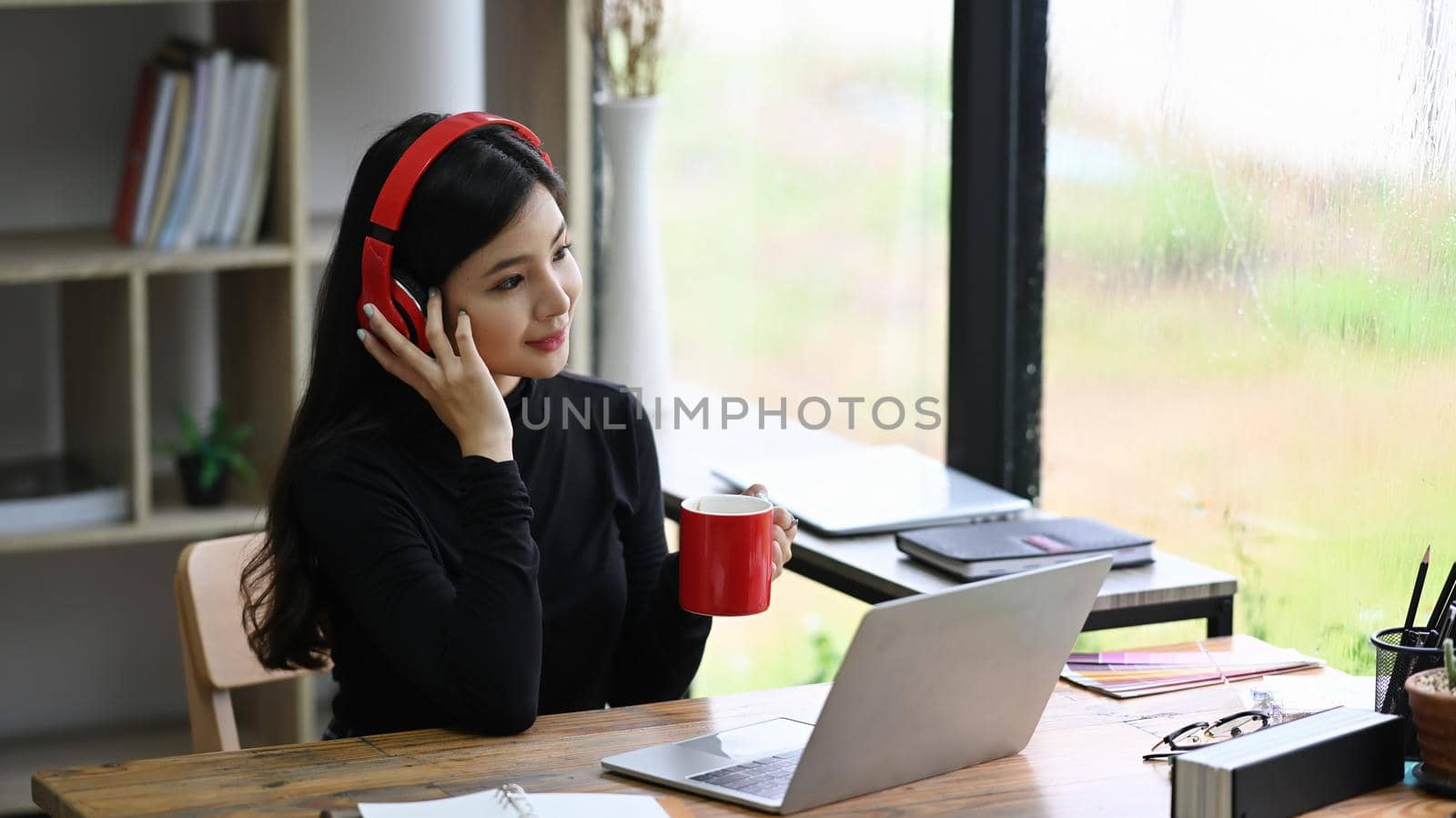 Attractive young woman designer wearing headphone and drinking coffee in office.