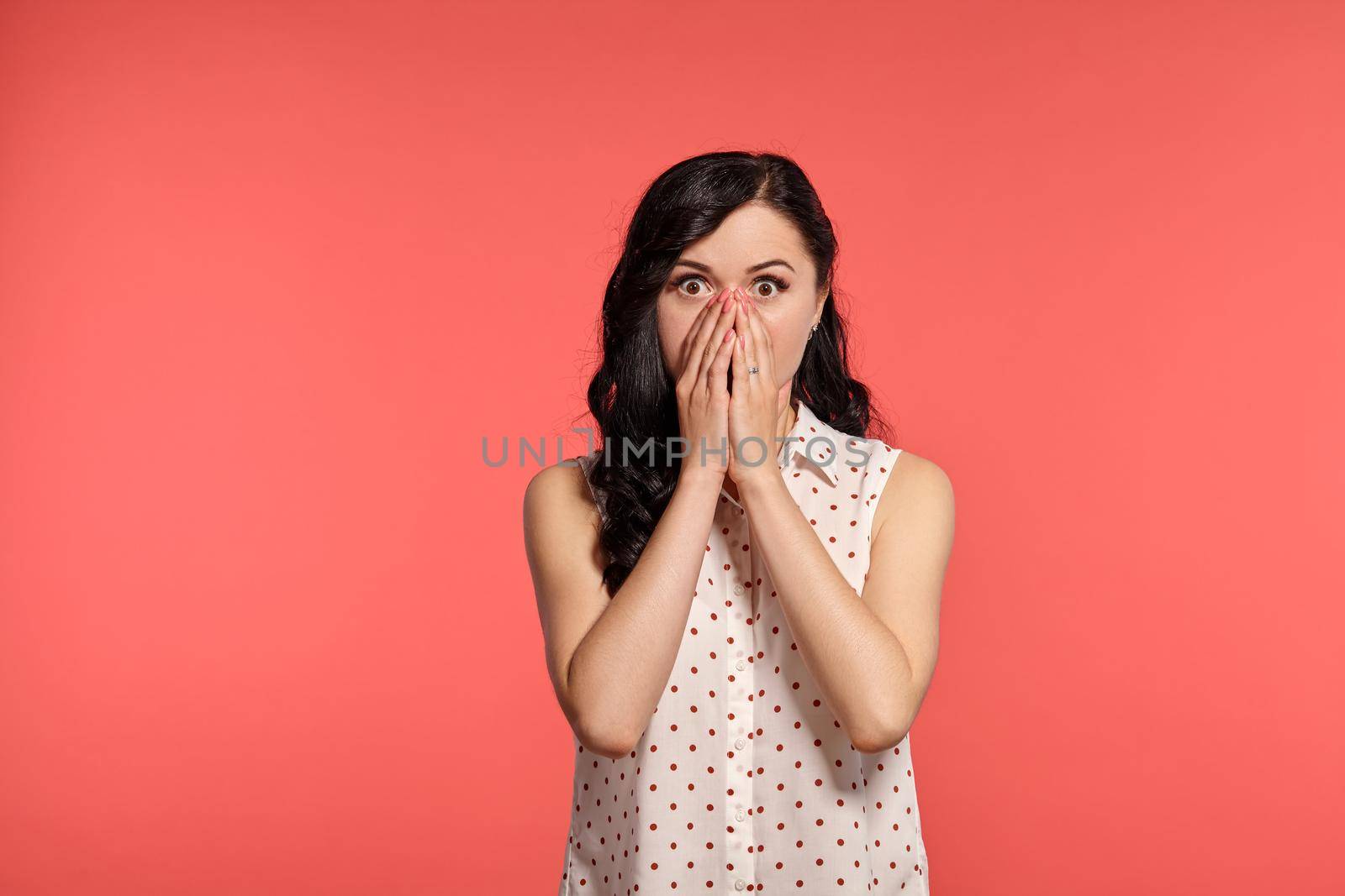 Studio shot of an elegant adolescent girl looking surprised, wearing casual white polka dot blouse. Little brunette female has closed her mouth with hands and looking at the camera, posing over a pink background. People and sincere emotions.