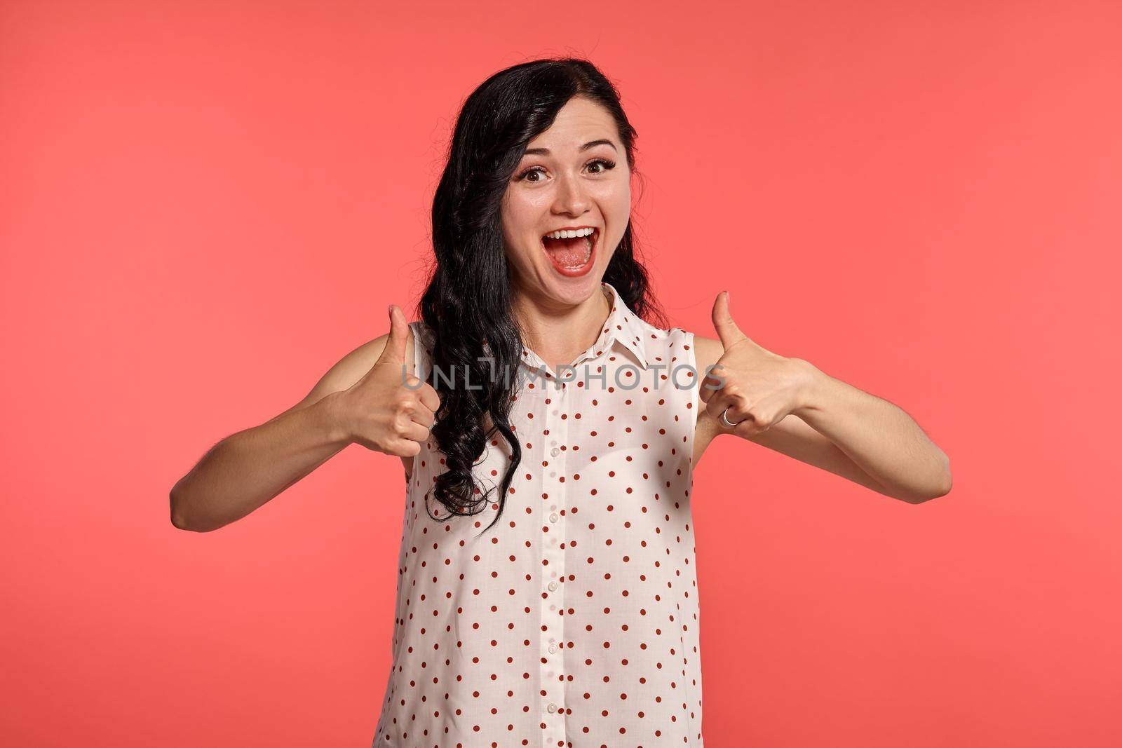 Studio shot of an amazing teen lady wearing casual white polka dot blouse. Little brunette female is showing thumbs up and smiling posing over a pink background. People and sincere emotions.