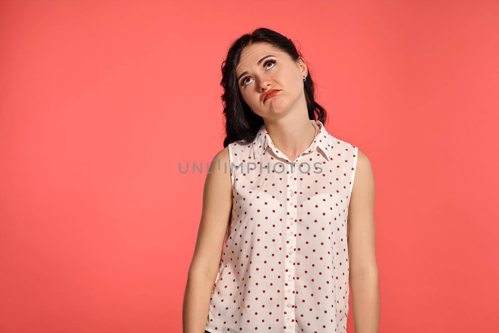 Studio shot of an adorable girl teenager looking unhappy, wearing casual white polka dot blouse. Little brunette female posing over a pink background. People and sincere emotions.