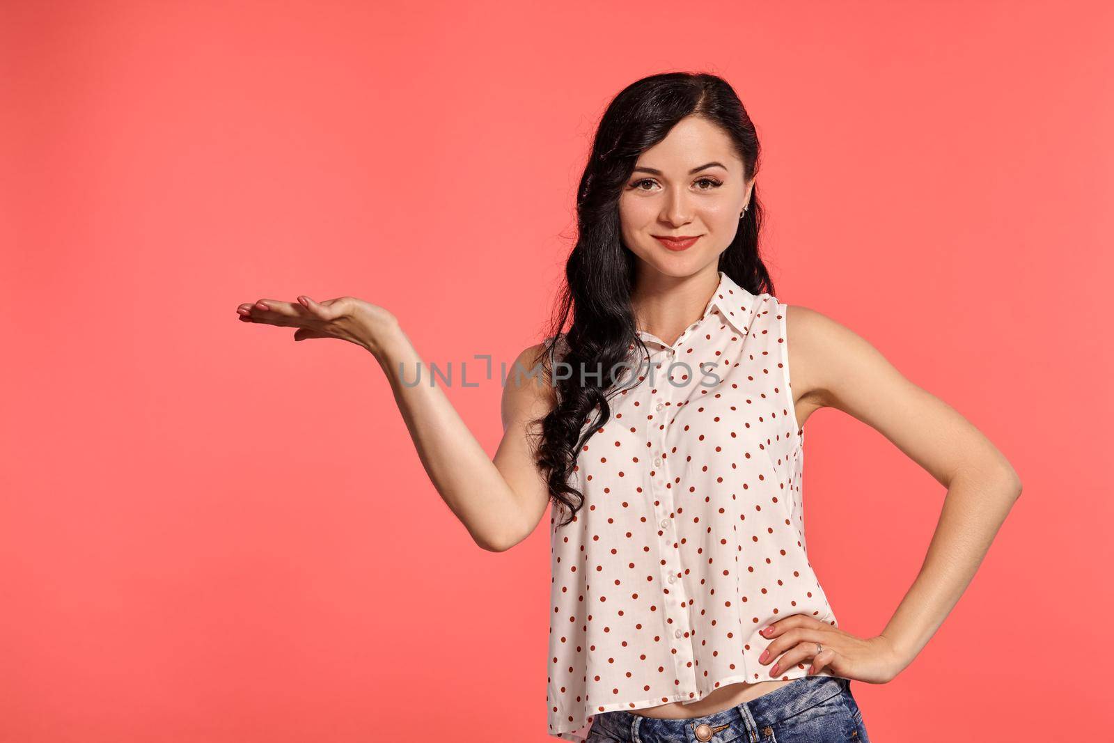 Close-up studio shot of a nice little woman, wearing casual white polka dot blouse and denim short shorts. Little brunette female acting like holding something in her arm, posing over a pink background. People and sincere emotions.