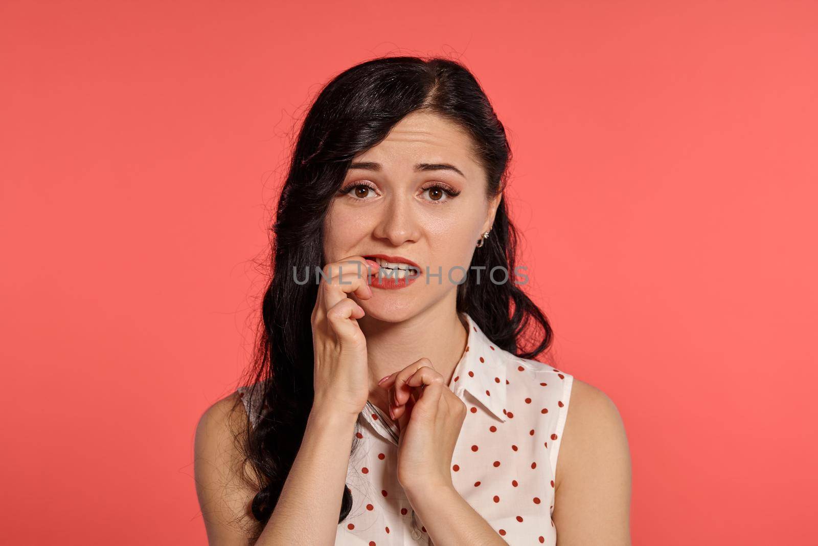 Close-up studio shot of a pretty little woman, wearing casual white polka dot blouse. Little brunette female looking confused, posing over a pink background. People and sincere emotions.