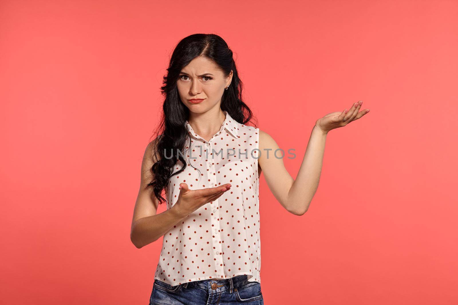 Studio shot of a lovely little woman, wearing casual white polka dot blouse and denim short shorts. Little brunette female looking upset, posing over a pink background. People and sincere emotions.