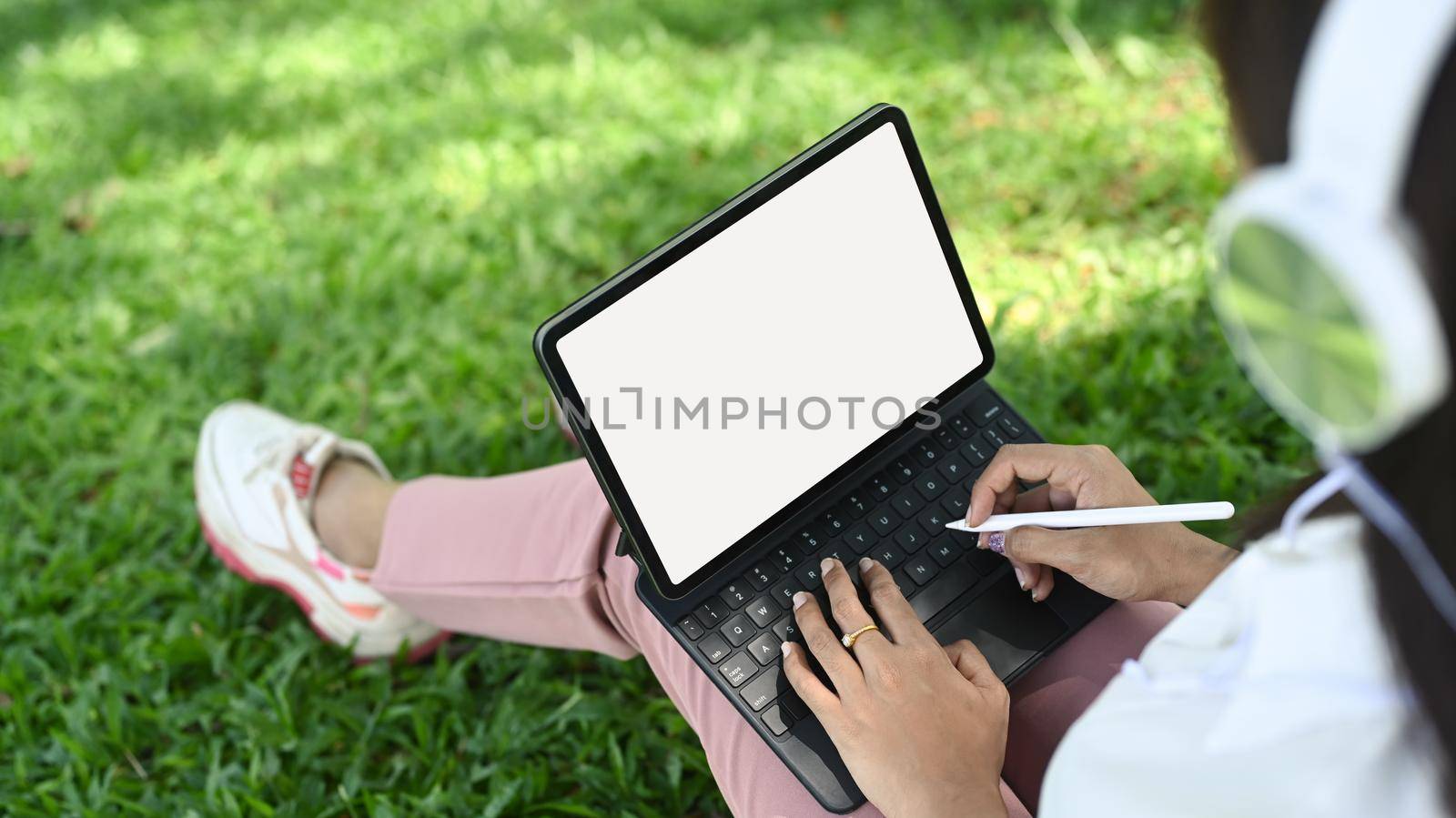 Young woman wearing head phone and working with digital tablet while sitting on green grass in the nature park.