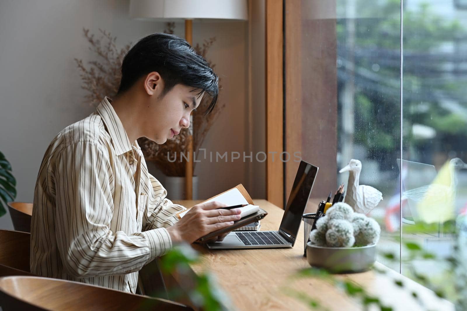 Side view businessman sitting in front of laptop computer and reading information on notebook.