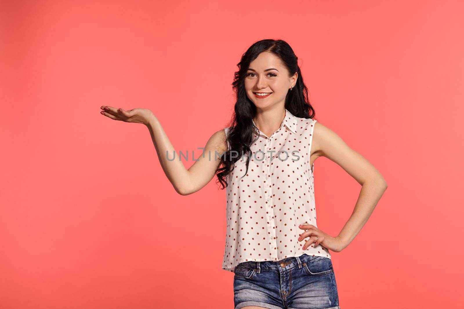 Close-up studio shot of a nice girl teenager looking happy, wearing casual white polka dot blouse and denim short shorts. Little brunette female acting like holding something in her hand, posing over a pink background. People and sincere emotions.