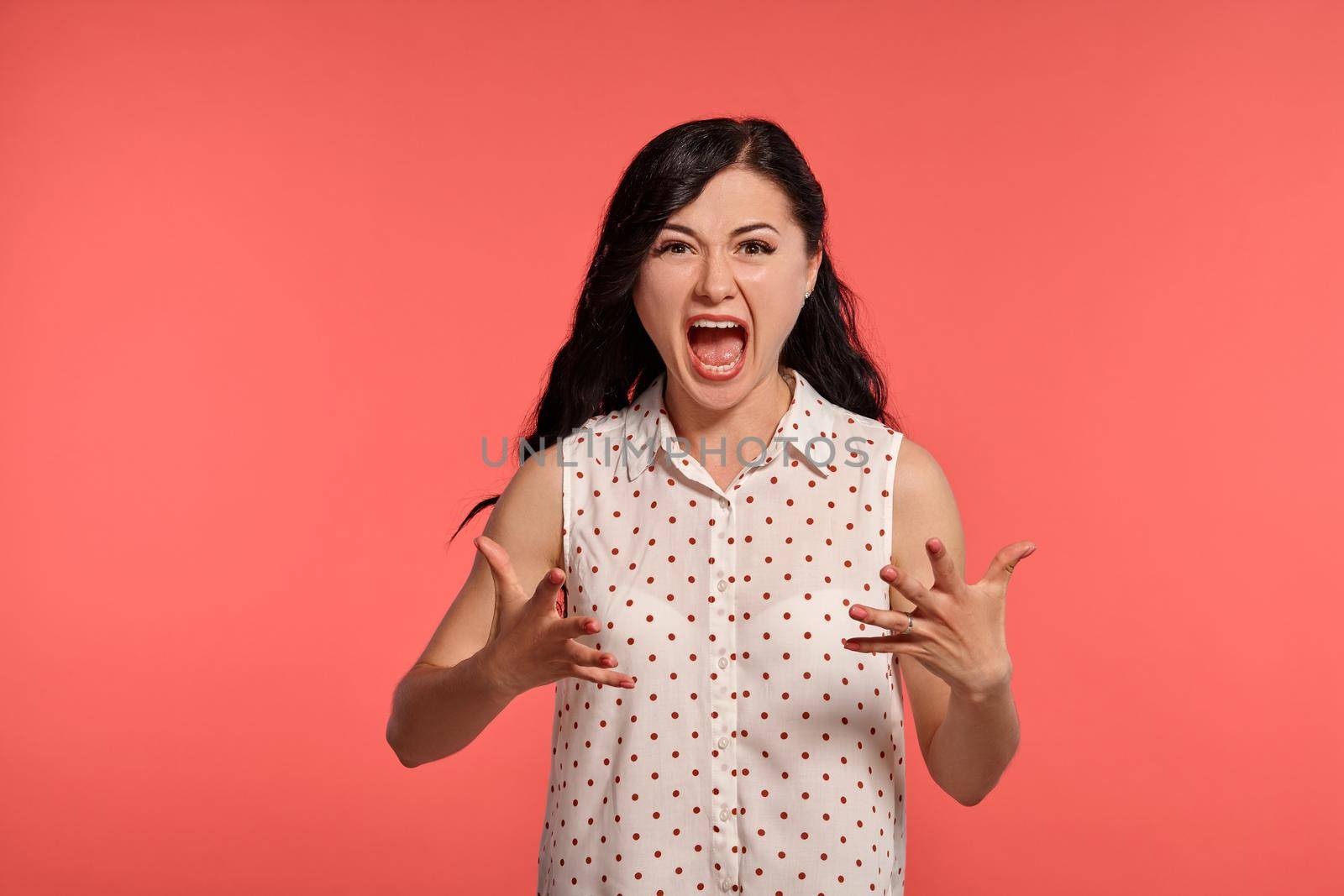 Studio shot of a beautiful girl teenager, screaming loudly, wearing casual white polka dot blouse. Little brunette female feeling unhappy, posing over a pink background. People and sincere emotions.