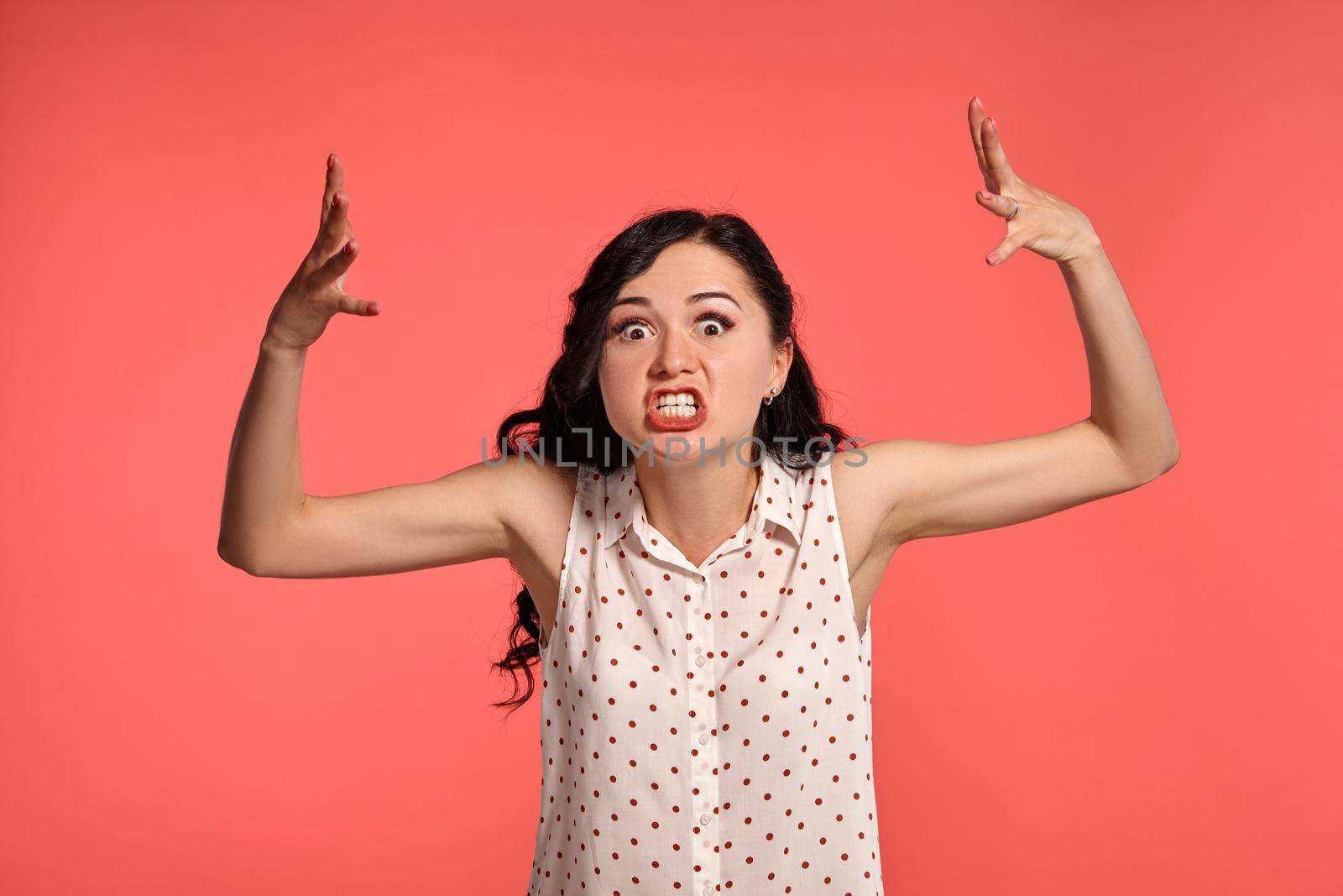 Studio shot of an adorable little woman looking angry, wearing casual white polka dot blouse. Little brunette female feeling unhappy, posing over a pink background. People and sincere emotions.