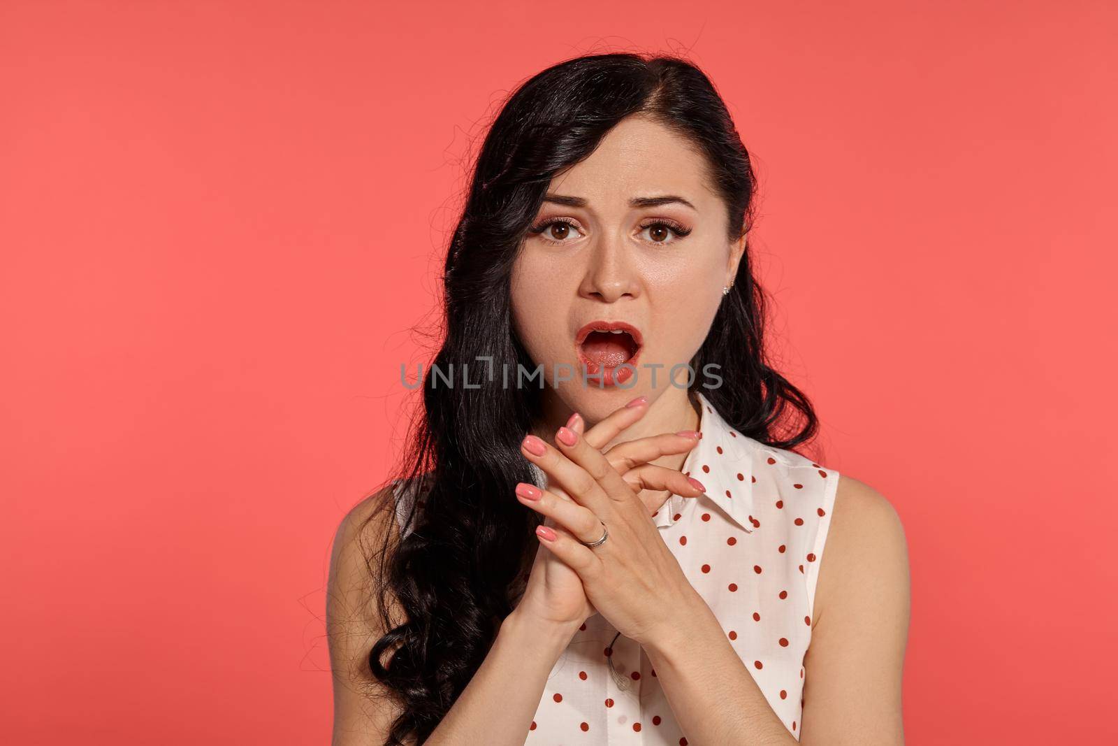 Close-up studio shot of a pretty teen lady, wearing casual white polka dot blouse. Little brunette female looking shocked posing over a pink background. People and sincere emotions.