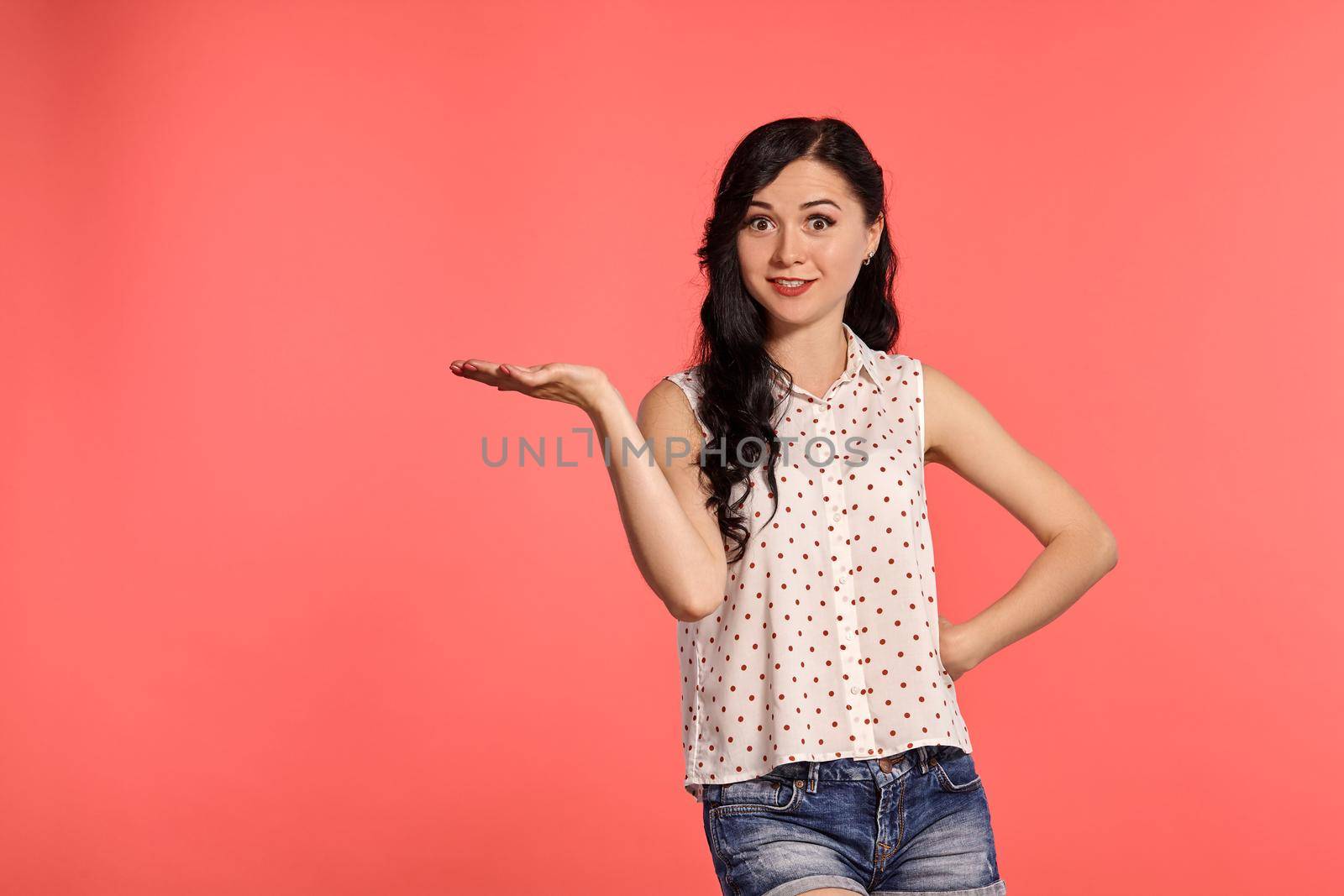 Close-up studio shot of a nice teen lady, wearing casual white polka dot blouse and denim short shorts. Little brunette female acting like holding something in her hand, posing over a pink background. People and sincere emotions.