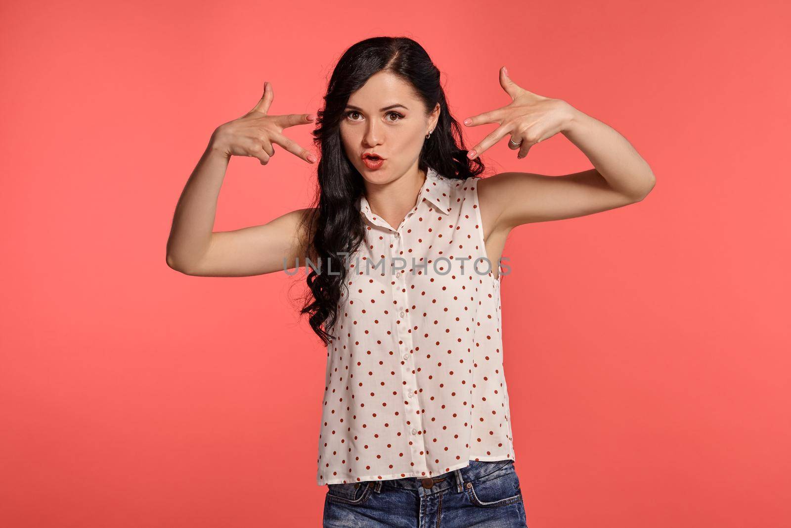 Studio shot of a lovely teen lady, wearing casual white polka dot blouse and denim short shorts. Little brunette female gesticulating posing over a pink background. People and sincere emotions.