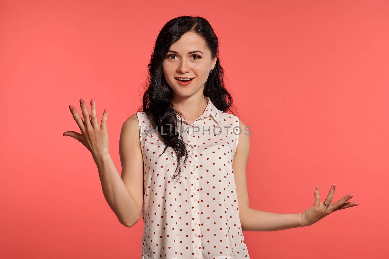 Studio shot of a charming little woman, wearing casual white polka dot blouse. Little brunette female looking wondered, posing over a pink background. People and sincere emotions.