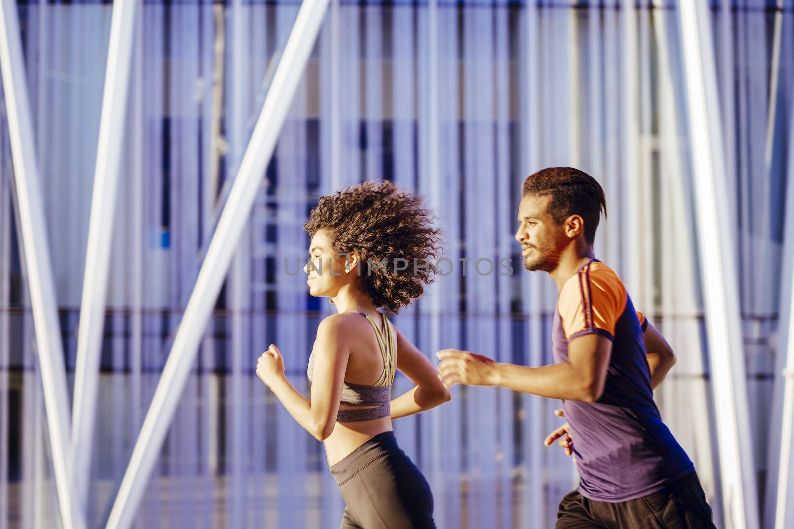 portrait of a beautiful latin woman and a sporty black man running together at sunset in the city street with buildings in the background. urban sports workout in couple and healthy lifestyle concept