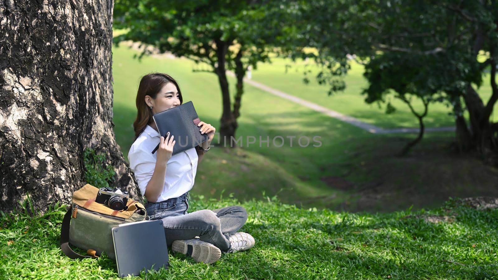 Peaceful young woman sitting under tree in the park and reading interesting book. by prathanchorruangsak