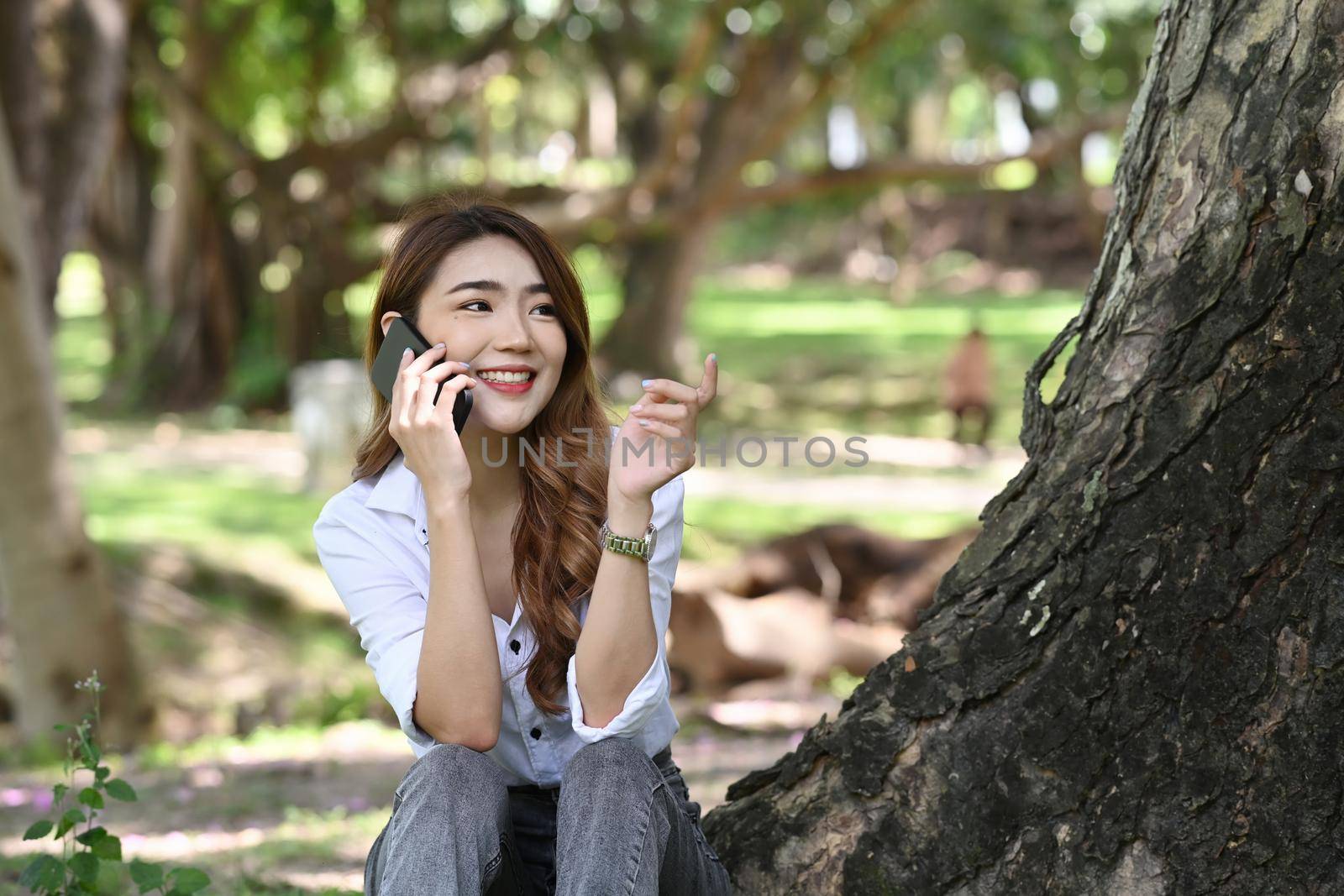 Smiling woman siting in public park and talking on mobile phone. by prathanchorruangsak