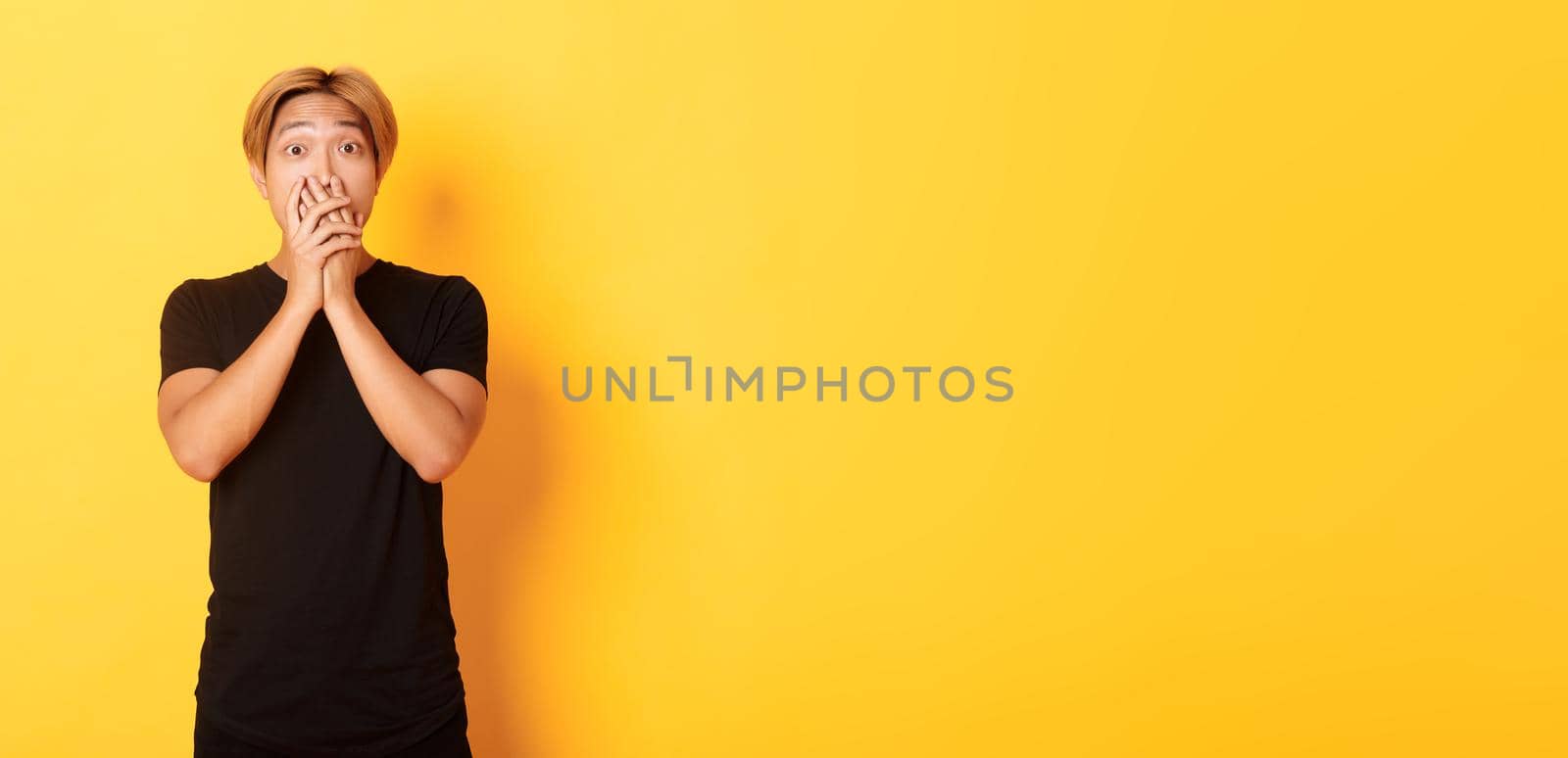 Portrait of surprised asian handsome guy with blond hair, cover mouth with hands and stare at camera impressed, yellow background by Benzoix
