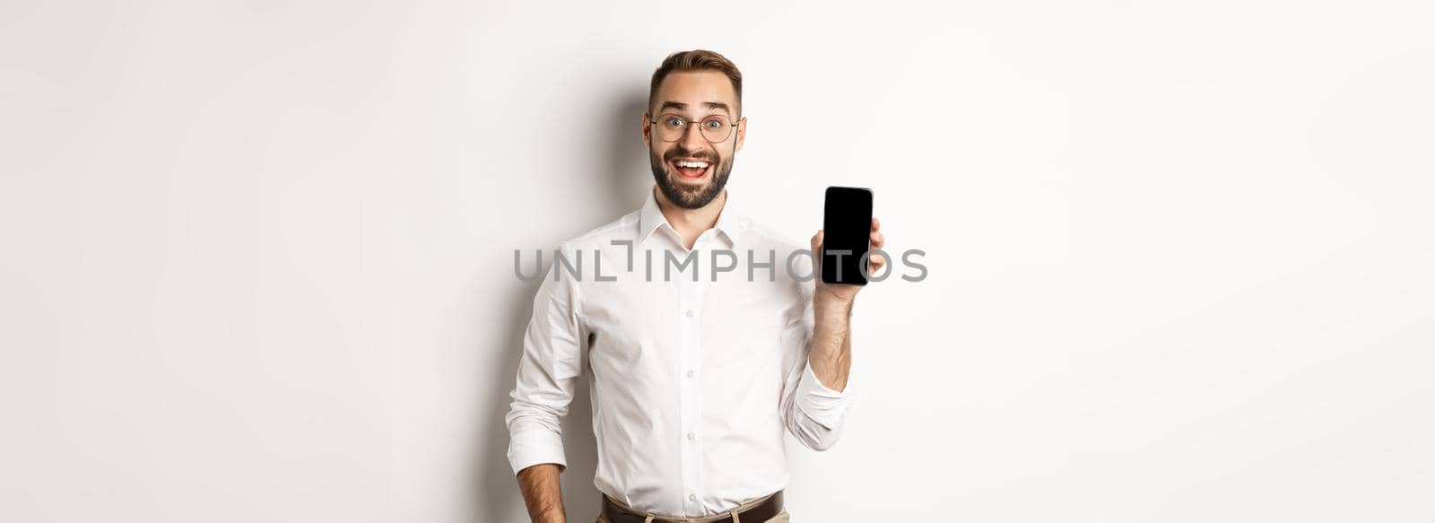 Amazed handsome businessman showing smartphone screen app, standing over white background.