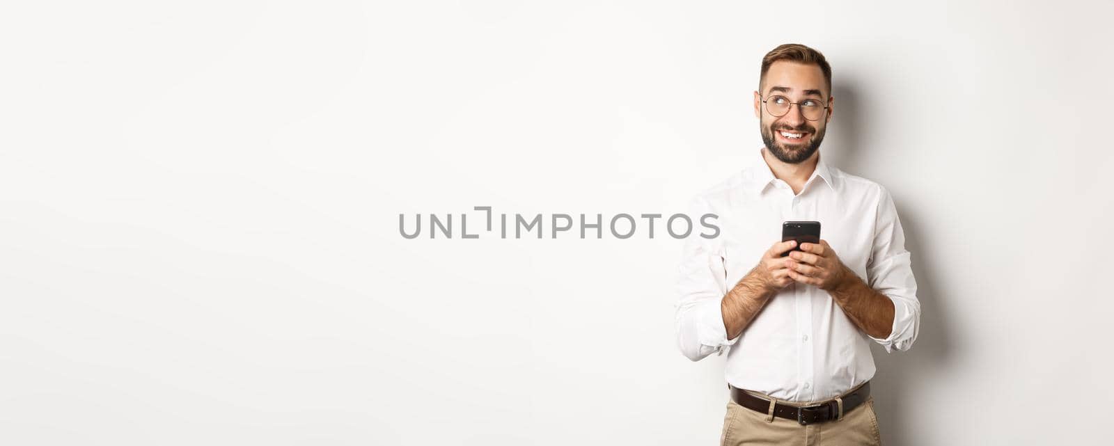 Thouthful handsome manager using mobile phone and thinking about answering message, looking at upper left corner and smiling, standing over white background by Benzoix