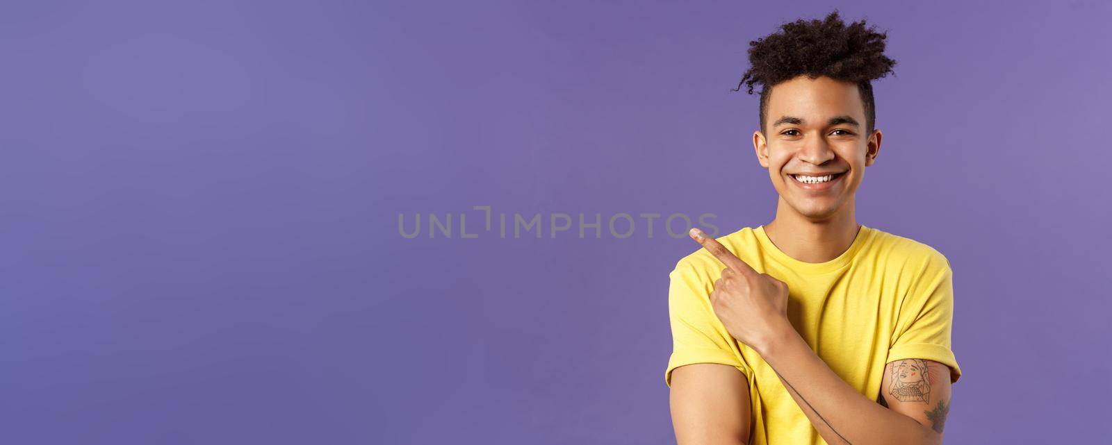 Close-up portrait of enthusiastic, happy young hipster male with dreads, beaming smile and pointing finger upper left corner, present cool product, introduce something really good, purple background.