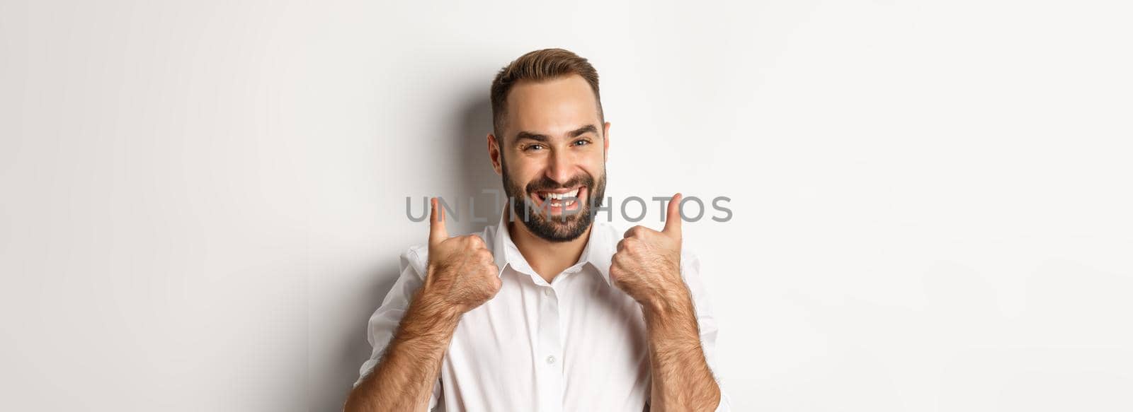 Close-up of satisfied bearded guy in white shirt, showing thumbs up in approval, like and agree, positive answer, white background.