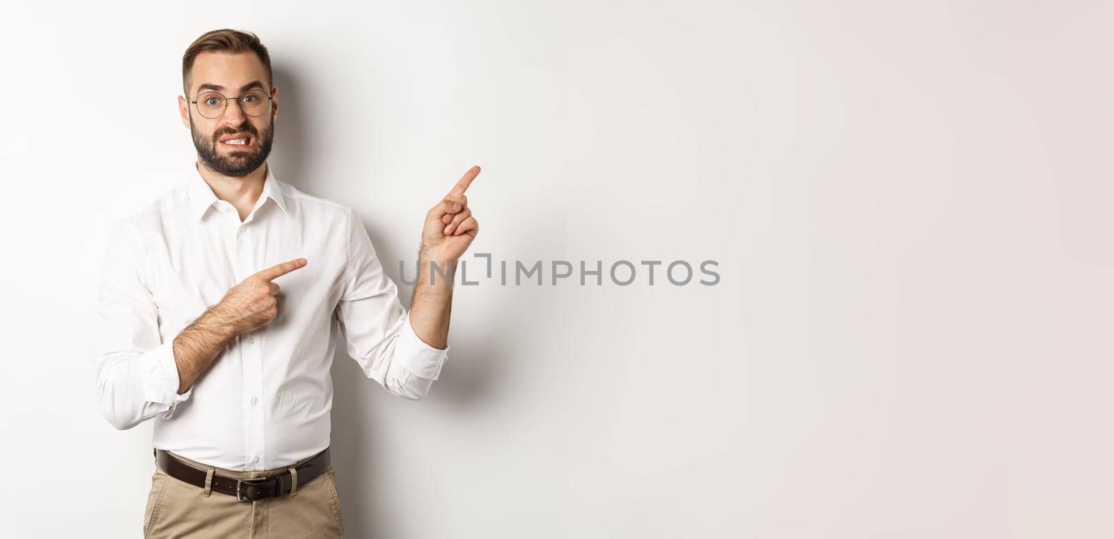 Doubtful and worried man looking indecisive, pointing at upper right corner hesitant, standing over white background.