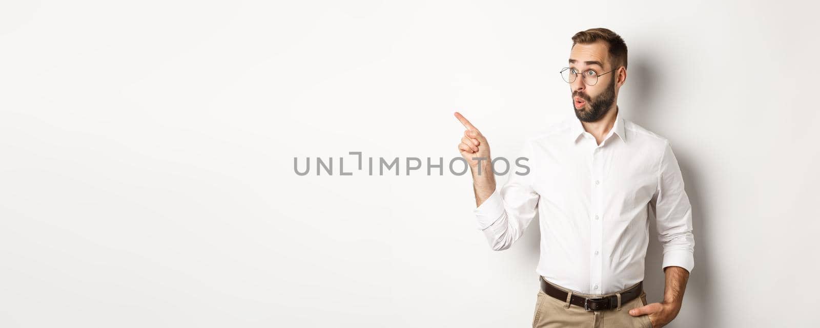 Intrigued young man in glasses pointing finger left, looking at promo offer interested, standing against white background.