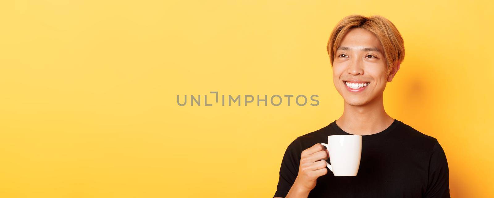 Close-up of happy handsome young asian guy with blond hair, looking dreamy and smiling while drinking coffee or tea, standing over yellow background by Benzoix