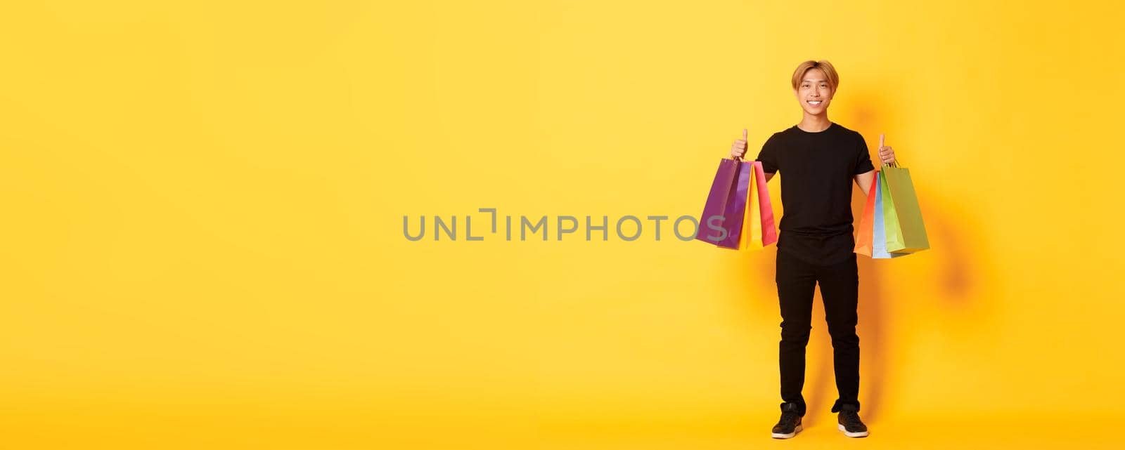 Full-length of attractive korean guy in black clothes, showing thumbs-up, holding shopping bags, yellow background.