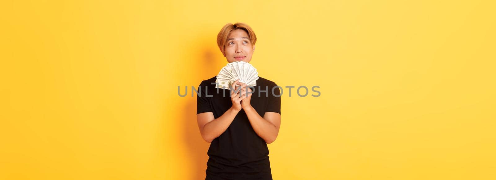 Portrait of dreamy handsome asian guy showing his savings and thinking, looking upper left corner, holding money, standing yellow background.