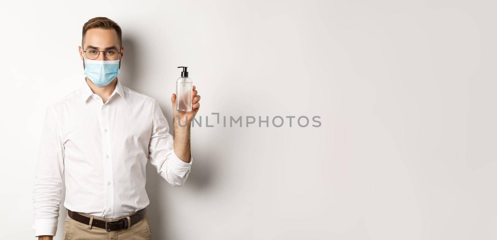 Covid-19, social distancing and quarantine concept. Employer in medical mask showing hand sanitizer, asking to use antiseptic at work, standing over white background.
