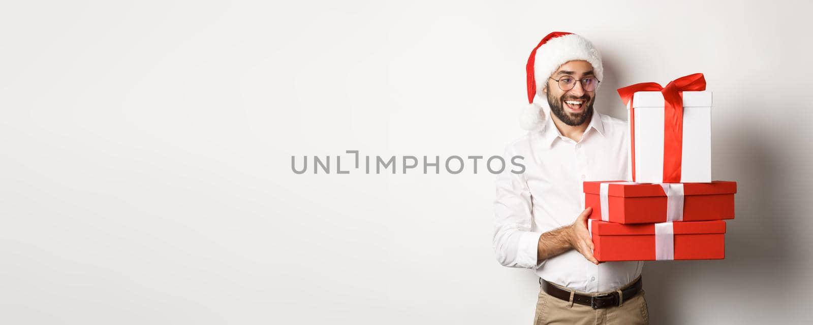 Winter holidays and celebration. Happy guy bring christmas presents, holding gifts and wearing Santa hat, standing over white background.